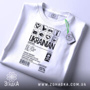 Футболка незламний українець, білого кольору, зображена з верхнього ракурсу, фон світло-сірий, принт чорного кольору з символами та текстом, на комірці є етикетка з зеленим шнурком.