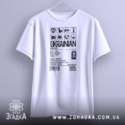 Футболка незламний українець, білого кольору, зображена з фронтального ракурсу на вішалці, фон світло-сірий, принт чорного кольору з символами та текстом, що підкреслює українську ідентичність.