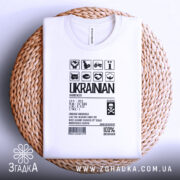 Футболка незламний українець, білого кольору, зображена зверху на плетеній кошику, фон білий, принт чорного кольору з символами та текстом, що підкреслює українську ідентичність.