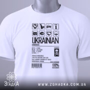 Футболка незламний українець, білого кольору, зображена з верхнього ракурсу, фон світло-сірий, принт чорного кольору з символами та текстом, на комірці є етикетка з зеленим шнурком.
