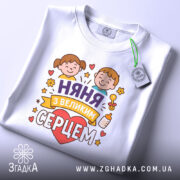 Футболка няня, білого кольору, зображена зверху, складена на поверхні, фон світло-сірий, принт з двома дітьми, серцем та написом 'няня з великим серцем', на комірці видна етикетка.