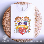 Футболка няня, білого кольору, зображена зверху, складена на плетеній основі, фон світлий, принт з двома дітьми, серцем та написом 'няня з великим серцем', на комірці видна етикетка.