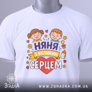 Футболка няня, білого кольору, зображена зверху, складена на поверхні, фон світло-сірий, принт з двома дітьми, серцем та написом 'няня з великим серцем', на комірці видна етикетка.