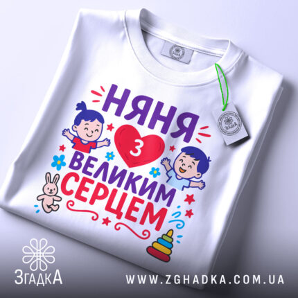 Футболка няня з серцем, білого кольору, зображена зверху на плоскій поверхні, фон світло-сірий, принт яскравий, містить малюнки дітей, серце, текст "НЯНЯ 3 ВЕЛИКИМ СЕРЦЕМ" та елементи декору, з біркою на комірці.