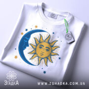 Футболка ніч і день, біла, зображена зверху, на фоні світло-сірого кольору, з принтом, що зображає сонце та місяць, з етикеткою зеленого кольору, сонце жовтого кольору з деталями, а місяць синій.