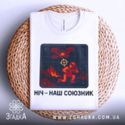 Футболка ніч наш союзник, білого кольору, зображена зверху на плетеній кошику, фон світло-сірий, принт з червоним і жовтим малюнком, що зображає людину в шоломі та дрон.