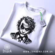 Футболка нічний бунтар, біла, складена на столі, з видимим етикеткою зеленого кольору, фон світло-сірий, принт з чорним малюнком обличчя персонажа з кучерявим волоссям і костюмом.