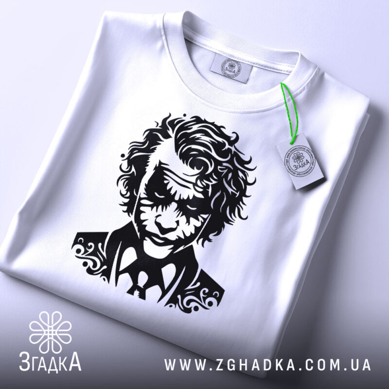 Футболка нічний бунтар, біла, складена на столі, з видимим етикеткою зеленого кольору, фон світло-сірий, принт з чорним малюнком обличчя персонажа з кучерявим волоссям і костюмом.