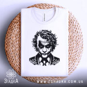 Футболка нічний бунтар, біла, складена на плетеній кошику, фон світло-сірий, принт з чорним малюнком обличчя персонажа з кучерявим волоссям і костюмом.