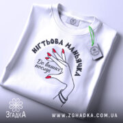 Футболка нігтьова маньячка, білого кольору, складена на поверхні, фон світло-сірий, принт з рукою з червоними нігтями та написом 'До ваших послуг', на комірці видна етикетка.