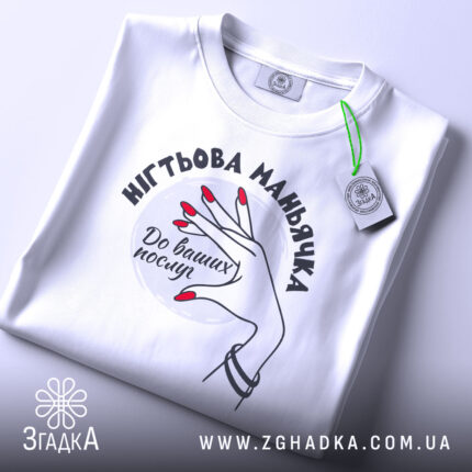 Футболка нігтьова маньячка, білого кольору, складена на поверхні, фон світло-сірий, принт з рукою з червоними нігтями та написом 'До ваших послуг', на комірці видна етикетка.