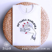 Футболка нігтьова маньячка, білого кольору, складена на плетеній кошику, фон світло-сірий, принт з рукою з червоними нігтями та написом 'До ваших послуг', видна етикетка на комірці.