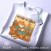 Футболка нікуди не спішу, білого кольору, складена на столі, фон світло-сірий, принт з котом у халаті, що сидить на балконі з чашкою в руках, поряд з ним маленьке котеня, на футболці є етикетка.