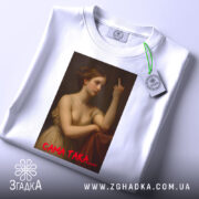 Футболка ніжність провокує, біла, зображена зверху, на фоні світлого кольору, принт з класичним зображенням жінки, що показує середній палець, з червоним написом 'САМА ТАКА....', з етикеткою зеленого кольору.