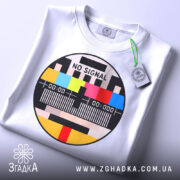 Футболка No Signal білого кольору, складена на столі, ракурс зверху, фон світло-сірий, принт у формі кола з написом 'NO SIGNAL' та кольоровими смужками, з етикеткою на зеленому шнурку.