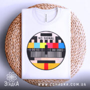 Футболка No Signal білого кольору, складена на плетеній кошику, ракурс зверху, фон білий, принт у формі кола з написом 'NO SIGNAL' та кольоровими смужками.