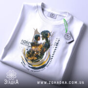 Футболка нове покоління, білого кольору, зображена зверху, на ній є етикетка, фон світлий, принт зображає роботизовану фігуру, що тримає дитину, з яскравими кольорами та абстрактними елементами.