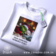 Футболка новорічний грінч білого кольору, складена на поверхні, фон світлий, принт з грінчем і собакою в новорічних шапках, текст "Щасливого Нового Року" під малюнком, на футболці є етикетка з зеленим шнурком.