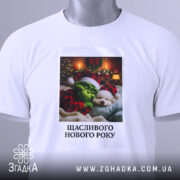 Футболка новорічний грінч білого кольору, складена на поверхні, фон світлий, принт з грінчем і собакою в новорічних шапках, текст "Щасливого Нового Року" під малюнком, на футболці є етикетка з зеленим шнурком.