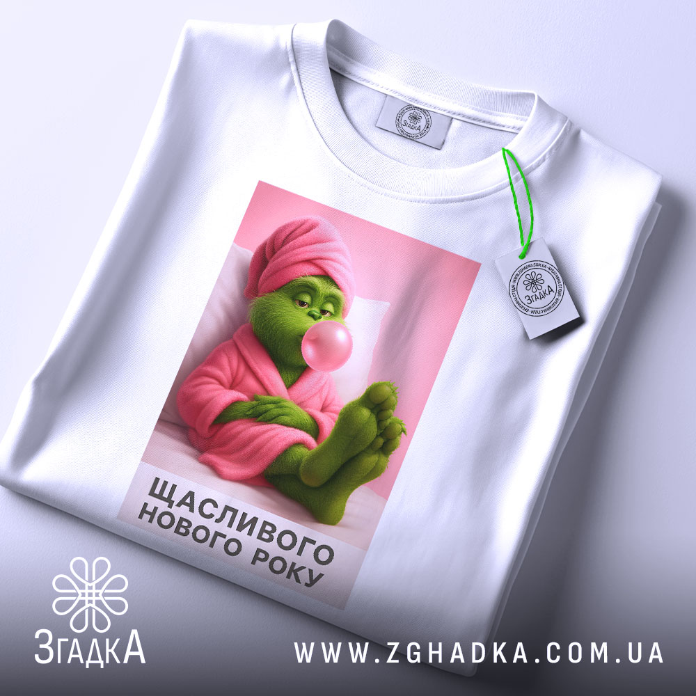Футболка новорічний грінч, біла, зображена з верхнього ракурсу, фон світло-сірий, принт з грінчем у рожевому халаті, який жує жувальну гумку, текст "Щасливого Нового Року" під малюнком, на футболці є етикетка.