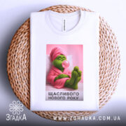 Футболка новорічний грінч, біла, зображена з верхнього ракурсу, фон світло-сірий, принт з грінчем у рожевому халаті, який жує жувальну гумку, текст "Щасливого Нового Року" під малюнком, футболка лежить на плетеній основі.