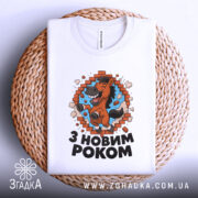 Футболка новорічний кінь, біла, складена на плетеній кошику, фон світло-сірий, принт з кінем, що стрибає через цегляну стіну, текст "З НОВИМ РОКОМ" під принтом.