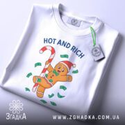 Футболка новорічний пряник білого кольору, складена на поверхні, фон світлий, принт з пряником у червоній шапці, що тримає цукерку, оточений зеленими купюрами та написом 'HOT AND RICH', на футболці є етикетка з логотипом.