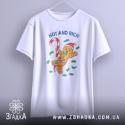 Футболка новорічний пряник білого кольору, підвішена на вішалці, фон світлий, принт з пряником у червоній шапці, що тримає цукерку, оточений зеленими купюрами та написом 'HOT AND RICH'.
