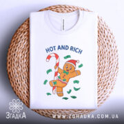 Футболка новорічний пряник білого кольору, складена на плетеній кошику, фон світлий, принт з пряником у червоній шапці, що тримає цукерку, оточений зеленими купюрами та написом 'HOT AND RICH'.