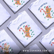 Футболка новорічний пряник білого кольору, зображена з фронтального ракурсу на манекені, фон світлий, принт з веселим пряником у червоній шапці, що тримає цукерку, оточений зеленими купюрами та написом 'HOT AND RICH'.