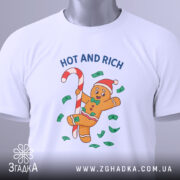 Футболка новорічний пряник білого кольору, складена на поверхні, фон світлий, принт з пряником у червоній шапці, що тримає цукерку, оточений зеленими купюрами та написом 'HOT AND RICH', на футболці є етикетка з логотипом.