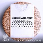 Футболка новий алфавіт, біла, складена на плетеній кошику, фон світло-сірий, принт чорний з літерами українського алфавіту.
