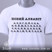 Футболка новий алфавіт, біла, складена на поверхні, фон світло-сірий, принт чорний з літерами українського алфавіту, з зеленим ярликом.