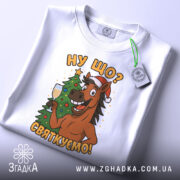 Футболка ну що святкуємо, біла, зображена зверху, на фоні світлого градієнту, принт з усміхненим конем у червоній шапці, що тримає келих з шампанським, поряд ялинка.