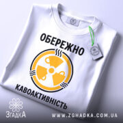 Футболка обережно кавоактивність, білого кольору, складена на поверхні, фон світло-сірий, принт жовтого кольору з символом радіації та текстом, на комірці видна етикетка.