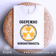 Футболка обережно кавоактивність, білого кольору, складена на плетеній кошику, фон світло-сірий, принт жовтого кольору з символом радіації та текстом, видно деталі тканини.