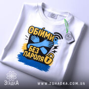 Футболка обійми без пароля, білого кольору, складена на поверхні, фон світло-сірий, принт з обіймами та текстом "Обійми без пароля" в жовтому, чорному та синьому кольорах, на футболці є етикетка з зеленим шнурком.