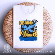 Футболка обійми без пароля, білого кольору, складена на плетеній кошику, фон світло-сірий, принт з обіймами та текстом "Обійми без пароля" в жовтому, чорному та синьому кольорах, видима етикетка на горловині.