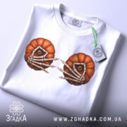 Футболка очі гарбуза, біла, складена на столі, фон світло-сірий, принт з двома гарбузами з очима та скелетними руками, на футболці є етикетка з зеленим шнурком.