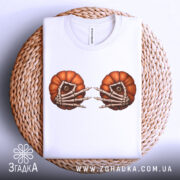 Футболка очі гарбуза, біла, складена на плетеній кошику, фон світло-сірий, принт з двома гарбузами з очима та скелетними руками, футболка акуратно розкладена.