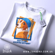 Футболка очі витріщив. Біла футболка з принтом собаки, зображена зверху, з етикеткою на шнурку, фон світло-сірий, принт кольоровий з текстом.