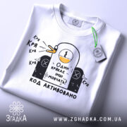 Футболка один крякає. Біла футболка на столі, з принтом качки, що крякає, та текстом "Один крякає – інші мовчать!". На футболці є етикетка з зеленим шнурком.