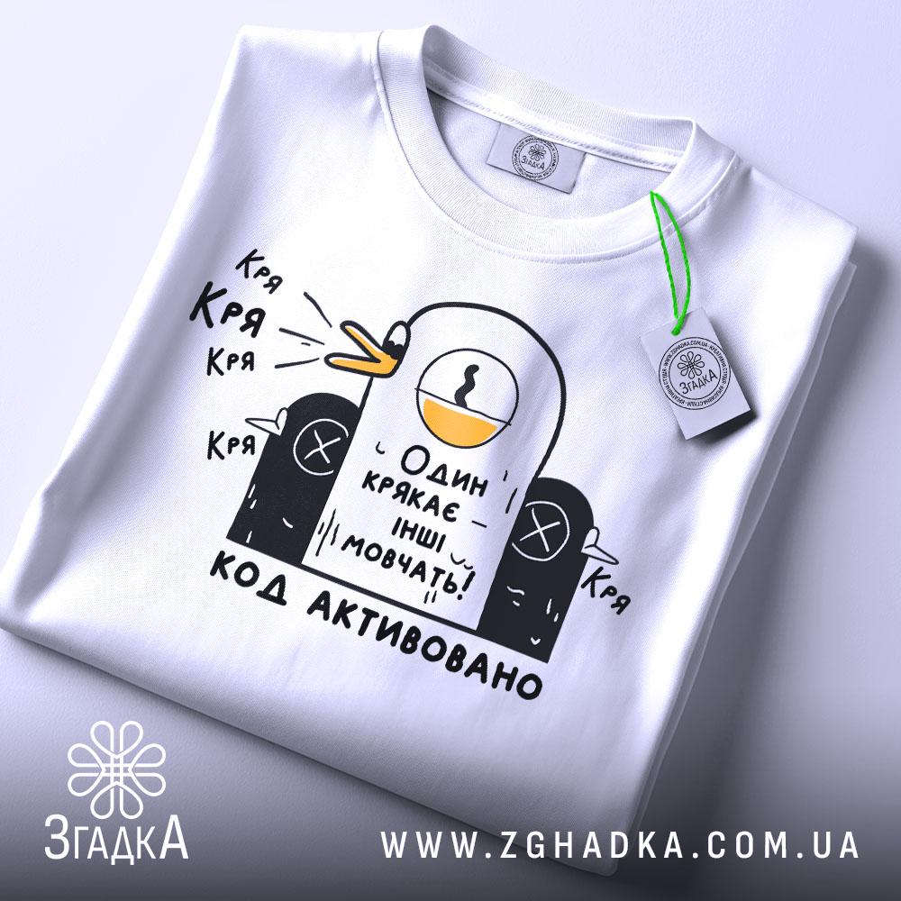 Футболка один крякає. Біла футболка на столі, з принтом качки, що крякає, та текстом "Один крякає – інші мовчать!". На футболці є етикетка з зеленим шнурком.