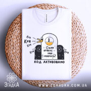 Футболка один крякає. Біла футболка, складена на плетеному кошику, з принтом качки, що крякає, та текстом "Один крякає – інші мовчать!". Фон світлий.