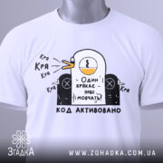 Футболка один крякає. Біла футболка на столі, з принтом качки, що крякає, та текстом "Один крякає – інші мовчать!". На футболці є етикетка з зеленим шнурком.