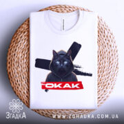 Футболка окак мем подарунок, складена біла футболка з принтом чорного кота в капюшоні, на плетеній кошику, на фоні світло-сірого кольору.