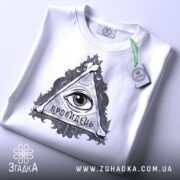 Футболка око провидця, біла, складена на поверхні, фон світло-сірий, принт у вигляді трикутника з оком та написом 'ПРОВИДЕЦЬ', на футболці є етикетка з зеленим шнурком.