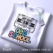 Футболка Old School білого кольору, складена на поверхні, з видимим етикеткою, фон світлий, принт з касетою та написом 'OLD SCHOOL' у яскравих кольорах.