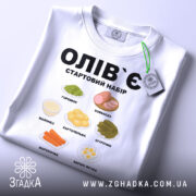 Футболка олів`є набір білого кольору, складена на поверхні, фон світло-сірий, принт містить малюнки овочів та інгредієнтів олів`є, текст чорного кольору: 'ОЛІВ`Є СТАРТОВИЙ НАБІР', на футболці є етикетка зеленого кольору.