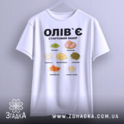 Футболка олів`є набір білого кольору, підвішена на вішалці, фон світло-сірий, принт містить малюнки овочів та інгредієнтів олів`є, текст чорного кольору: 'ОЛІВ`Є СТАРТОВИЙ НАБІР'.